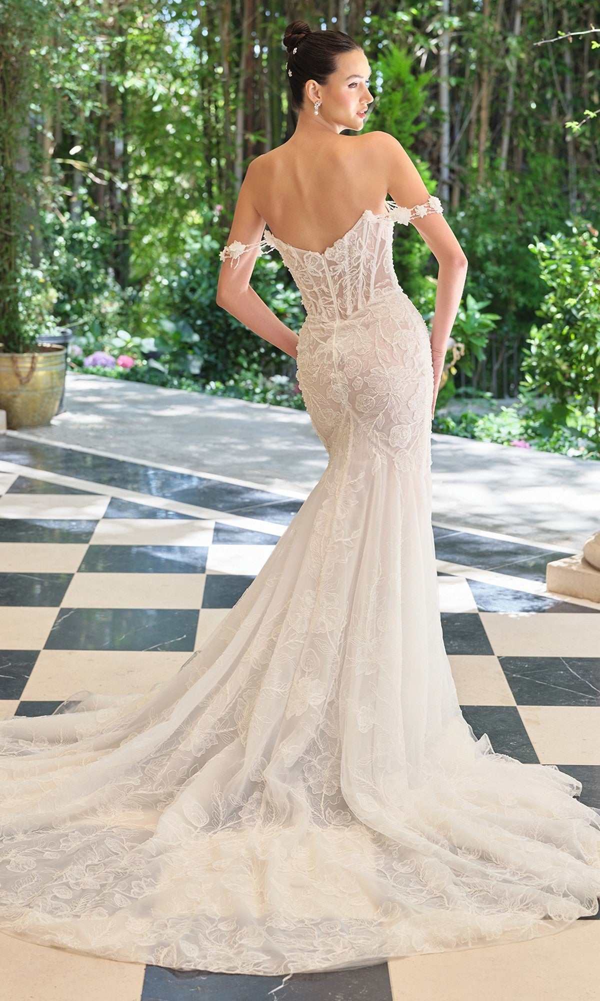 A woman stands on a black-and-white checkered floor in a picturesque garden, wearing the Andrea and Leo WL099 Off-White Wedding Dressโan off-the-shoulder mermaid gown with floral lace and a long train.